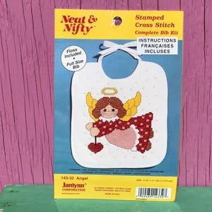 May include: A cross-stitch kit for a baby bib featuring an angel design. The bib is white with a cross-stitched angel in red, pink, yellow, and brown. The kit includes floss, a needle, and instructions. The bib measures 24.1 cm x 27.9 cm.