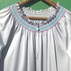 May include: A light gray vintage robe with a smocked yoke featuring blue and purple embroidery. The robe has a ruffled neckline and sleeves, and a button-down front. The robe is hanging on a wooden hanger.