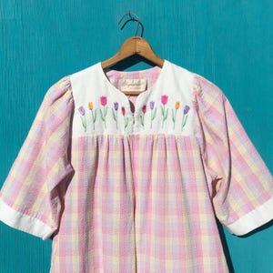 May include: A short-sleeved blouse featuring a pink, yellow, and white plaid pattern with a white yoke. The yoke is embroidered with a row of colorful tulips in shades of purple, pink, orange, and red. The garment includes a white collar and cuffs.