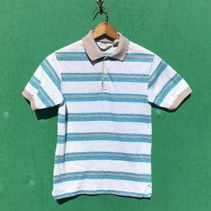 May include: A short-sleeve polo shirt featuring a white base with horizontal stripes in shades of blue and grey. The shirt has a beige collar and sleeve cuffs, a three-button placket, and a label inside the collar that reads Oakton.