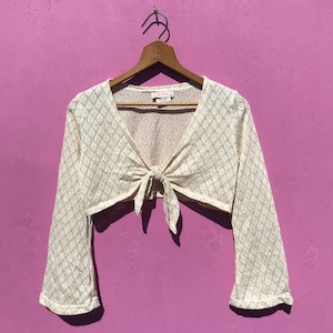 May include: Cream-colored, open-front bolero with long sleeves, hanging on a wooden hanger. The bolero features a diamond-patterned knit design and a tie closure at the waist. The background is a solid, bright pink.