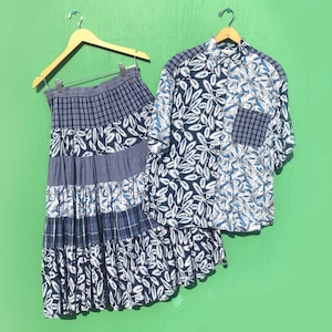 May include: A two-piece set of clothing, including a skirt and a button-down shirt, hanging against a green background. The skirt is tiered with blue and white leaf and plaid patterns. The shirt has a similar leaf pattern and a plaid pocket.