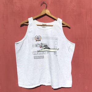 May include: A heather gray tank top featuring a detailed illustration of a yacht with the text "GIG HARBOR WASHINGTON". The graphic includes a nautical emblem and a depiction of a lighthouse. The tank top is hanging on a wooden hanger.