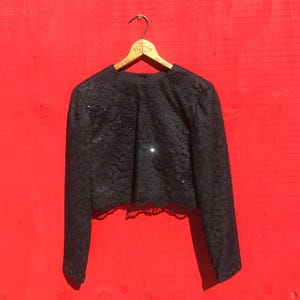 May include: A black lace bolero jacket with long sleeves hangs on a wooden hanger against a red background. The jacket features a round neckline and is embellished with sequins. The cropped design adds a touch of elegance.