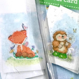 May include: Two small, illustrated cards featuring woodland creatures. One shows an orange fox with a butterfly, the other a hedgehog holding a dandelion. Both are set against a light blue background with floral accents.