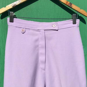 May include: High-waisted lavender trousers featuring a structured waistband with two small, round, light pink buttons. The pants have a clean, smooth finish and a central vertical seam running down the front, suspended from a metal clothing hanger.