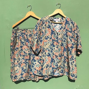 May include: A two-piece vintage outfit featuring a short-sleeved top and a skirt, both with a blue background and a floral pattern in shades of peach, teal, and cream. The top has a V-neck and the skirt has an elastic waistband. The outfit hangs on wooden hangers.