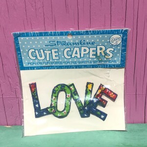 May include: A Streamline Cute Capers iron-on transfer package containing the word LOVE in fabric letters. The letters feature a multi-colored floral and leaf pattern in shades of green, blue, red, and yellow, enclosed in a clear plastic bag.