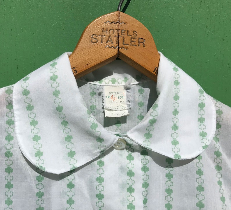 Puede incluir: Camisa blanca con cuello y estampado de tr&eacute;bol verde. La camisa tiene un cuello de Peter Pan y cierre de bot&oacute;n. La etiqueta dice "Official Irish School Size 12".