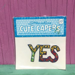 May include: A fabric iron-on patch featuring the word YES in a floral pattern. The Y is green with yellow flowers, while the E and S are red and yellow with blue flowers. The patch is in a blue and white polka dot package labeled Streamline CUTE CAPERS.