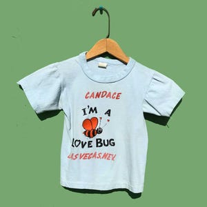 May include: Light blue children's t-shirt with the text "CANDACE I'M A LOVE BUG LAS VEGAS, NEV." and a cartoon bee graphic. The bee is orange and black with hearts. The shirt has short sleeves and is hanging on a wooden hanger.