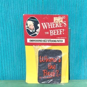 May include: A rectangular dark blue embroidered self-sticking patch featuring the orange text Where's the Beef? inside a clear plastic bag attached to a red and yellow cardboard backing card with an image of a person on a telephone.