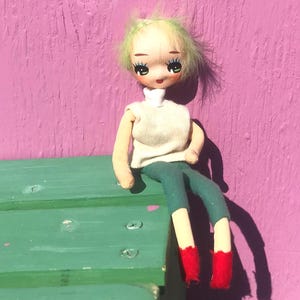 May include: A vintage doll with a painted face, green hair, and a white sweater. The doll is wearing teal pants and red shoes. It is sitting on a green surface against a pink background.