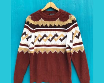 May include: A brown, white, and gold patterned sweater with a crew neck and long sleeves. The sweater features geometric designs and is hanging on a wooden hanger against a blue background. The sweater's design includes zig-zag and diamond patterns.