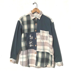 May include: A long-sleeved patchwork shirt with a mix of plaid and solid panels in shades of green, beige, and navy blue. The shirt features embroidered floral details and wooden buttons. The shirt is hanging on a wooden hanger.