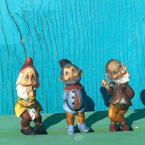 May include: Three ceramic garden gnomes with detailed features. One gnome wears a red hat and yellow coat, another has a blue coat, and the third has a brown coat. They stand against a turquoise wooden background.