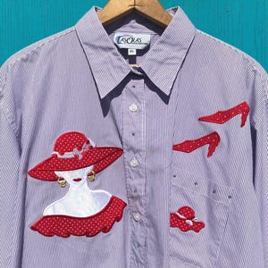 May include: A button-down shirt with a purple and white striped pattern. The shirt features appliqued red hats and high heels, along with a woman's face wearing a red hat. The shirt has a pocket on the left side and is hanging on a wooden hanger.
