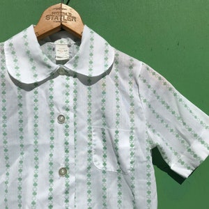 Puede incluir: Camisa blanca de manga corta con un estampado de tr&eacute;bol verde y un cuello Peter Pan. La camisa tiene un cierre de botones en la parte delantera y un bolsillo en el lado izquierdo.