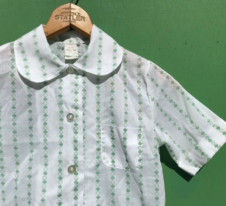 Puede incluir: Camisa blanca de manga corta con un estampado de tr&eacute;bol verde y un cuello Peter Pan. La camisa tiene un cierre de botones en la parte delantera y un bolsillo en el lado izquierdo.