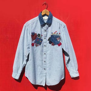 May include: A light blue striped button-down shirt with a denim collar. The shirt features colorful patchwork appliques of hearts, flowers, and gift boxes. The shirt is hanging on a wooden hanger against a red background.