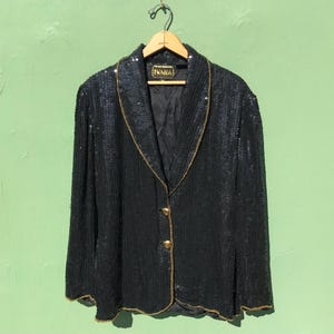 May include: A vintage black sequin blazer with gold trim and buttons. The blazer has a notched collar and long sleeves. The label reads "IVANA". The jacket is hanging on a wooden hanger against a green background.