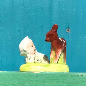 May include: A vintage ceramic figurine featuring a fawn and a child. The fawn is brown with white spots, and the child has white hair and a black and white outfit. The base is light green. The figurine is set against a blue background.