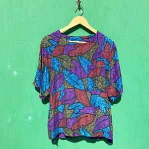 May include: A short-sleeved shirt featuring a vibrant, all-over leaf pattern in shades of purple, bright blue, and muted olive green. The garment includes a round neckline, raglan sleeves, and a single chest pocket on the left side, all displayed on a wooden hanger.