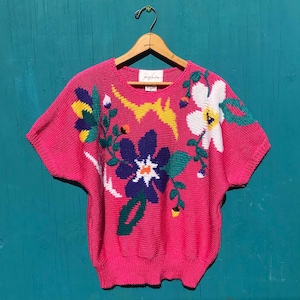 May include: A bright pink, short-sleeved sweater with a floral design. The flowers are in shades of white, blue, yellow, and green. The sweater is hanging on a wooden hanger against a teal background.