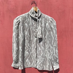 May include: A gray and white patterned blouse with a tie neck and long sleeves. The blouse is hanging on a wooden hanger against a red background.
