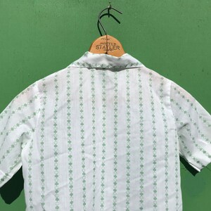 Puede incluir: Camisa blanca de manga corta con un patr&oacute;n de tr&eacute;bol verde. La camisa tiene un cuello abotonado y un dise&ntilde;o a rayas.