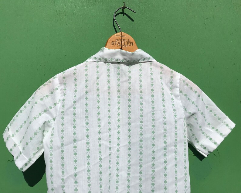 Puede incluir: Camisa blanca de manga corta con un patr&oacute;n de tr&eacute;bol verde. La camisa tiene un cuello abotonado y un dise&ntilde;o a rayas.