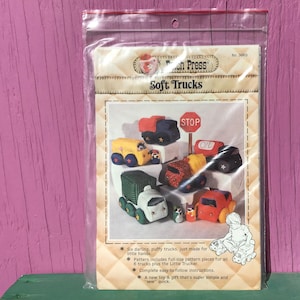 May include: A package for a sewing pattern titled "Soft Trucks" featuring illustrations of colorful, stuffed toy trucks. The package includes text that says "Six darling, puffy trucks, just made for little hands."