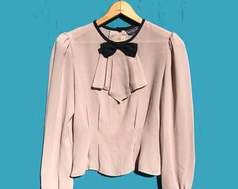 May include: A light pink long-sleeved blouse featuring a black bow at the neckline, black trim around the collar, and black cuffs. The garment has a pleated front panel and is displayed on a wooden hanger against a solid blue background.