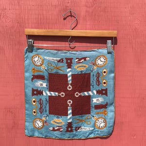 May include: A square, light blue silk scarf with a barber shop theme. The design includes clocks, scissors, barber poles, and other barber-related imagery in brown, white, and gold. The scarf is hanging on a wooden hanger.