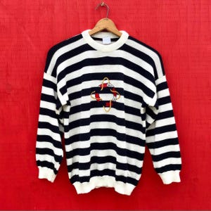 May include: A vintage nautical-themed sweater with horizontal navy blue and white stripes. The sweater features a crew neck, long sleeves, and a red and gold embroidered life preserver design on the front. The cuffs and collar are white.