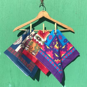 May include: Three square cotton bandanas hanging from a wooden clothes hanger by small wooden clips. The left bandana is dark blue with geometric patterns. The center one is red with cactus and snake motifs. The right one is blue with purple and pink geometric shapes.