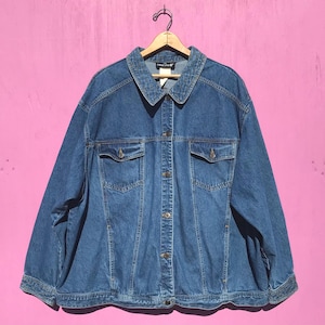 May include: A medium-wash denim jacket with a collared neckline and button-down closure. The jacket has two chest pockets and two side pockets. It is hanging on a wooden hanger against a pink background.