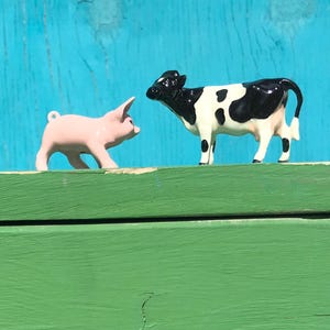 May include: A small pink ceramic pig figurine with a loop on its back and a black and white spotted ceramic cow figurine standing side by side. Both animal figures have a glossy finish and are positioned facing each other on a flat surface.