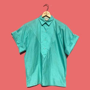 May include: A turquoise short-sleeve shirt with a collared neckline and a button-down front. The shirt has a unique V-shaped design on the front and is hanging on a wooden hanger. The background is a solid coral color.