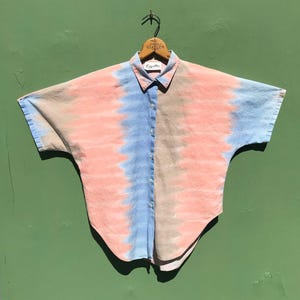 May include: A short-sleeved button-down shirt with a tie-dye pattern in shades of pink, blue, and tan. The shirt is hanging on a wooden hanger. The collar is light pink and the shirt has a relaxed fit.