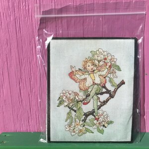 May include: Cross-stitch embroidery depicting a fairy with pink wings and a yellow dress, perched on a branch of a flowering tree with white blossoms.