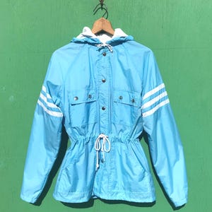 May include: A light blue vintage jacket with a white hood and white stripes on the sleeves. The jacket has a drawstring waist, two front pockets, and a button-up closure. The jacket is hanging on a wooden hanger.