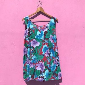 May include: A sleeveless top with a vibrant floral pattern. The top features large blue and purple flowers with green leaves against a red background. It hangs from a wooden hanger against a pink wall.