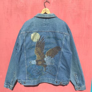May include: A light blue denim jacket featuring a painted eagle design on the back. The eagle is shown with outstretched wings against a night sky with a full moon. The jacket has a classic collar, button closures, and pockets.