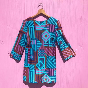 May include: A vintage, long-sleeve dress with a geometric pattern in teal, purple, and brown. The dress is hanging on a wooden hanger against a pink background. The pattern includes squares, rectangles, and diamond shapes.