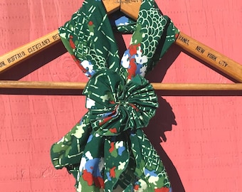 May include: A green scarf with a floral pattern in red, white, and blue is draped over a wooden hanger. The scarf is knotted in the center, creating a flower-like detail. The hanger has the words "HOTELS STATLER" and "BUFFALO, CLEVELAND, DE" printed on it.