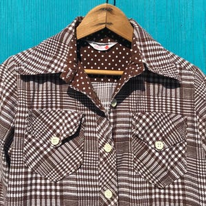 May include: A brown and white plaid button-up shirt with two front pockets. The collar and inner lining feature a brown polka dot pattern. The shirt is hanging on a wooden hanger.