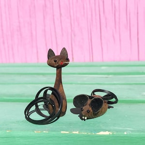 May include: Wooden cat and mouse figurines. The cat is tall and slender with a long neck, black tail, and a red nose. The mouse is small and lying down with black ears. Both are brown and sit on a green surface with a pink background.