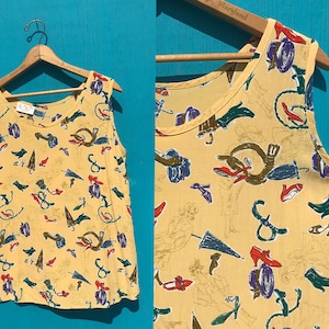 May include: A sleeveless, yellow top with a repeating pattern of umbrellas, shoes, hats, and other accessories. The top is hanging on a wooden hanger against a blue background. The print includes red, blue, green, and purple accents.