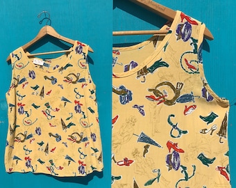 May include: A sleeveless, yellow top with a repeating pattern of umbrellas, shoes, hats, and other accessories. The top is hanging on a wooden hanger against a blue background. The print includes red, blue, green, and purple accents.
